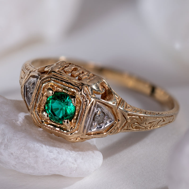 Late Art Deco 14K yellow gold ring featuring a vivid round green emerald and diamond accents displayed on white marble stone.