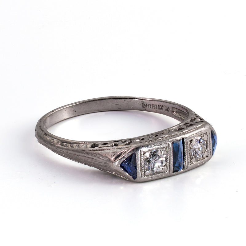 Left-side profile perspective of a geometric 1930s Art Deco ring, showcasing the engraved shoulders and the precise setting of the old-cut diamond and triangular blue sapphire.