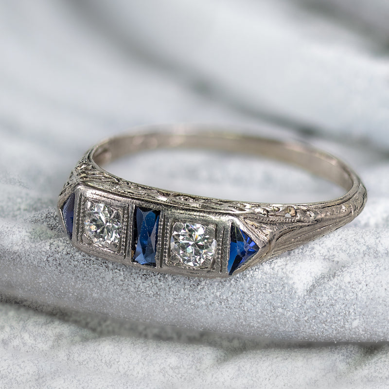 An angled hero view of an authentic Art Deco platinum ring, featuring two hand-cut Old European diamonds and geometric deep blue sapphire accents, popular circa 1930.